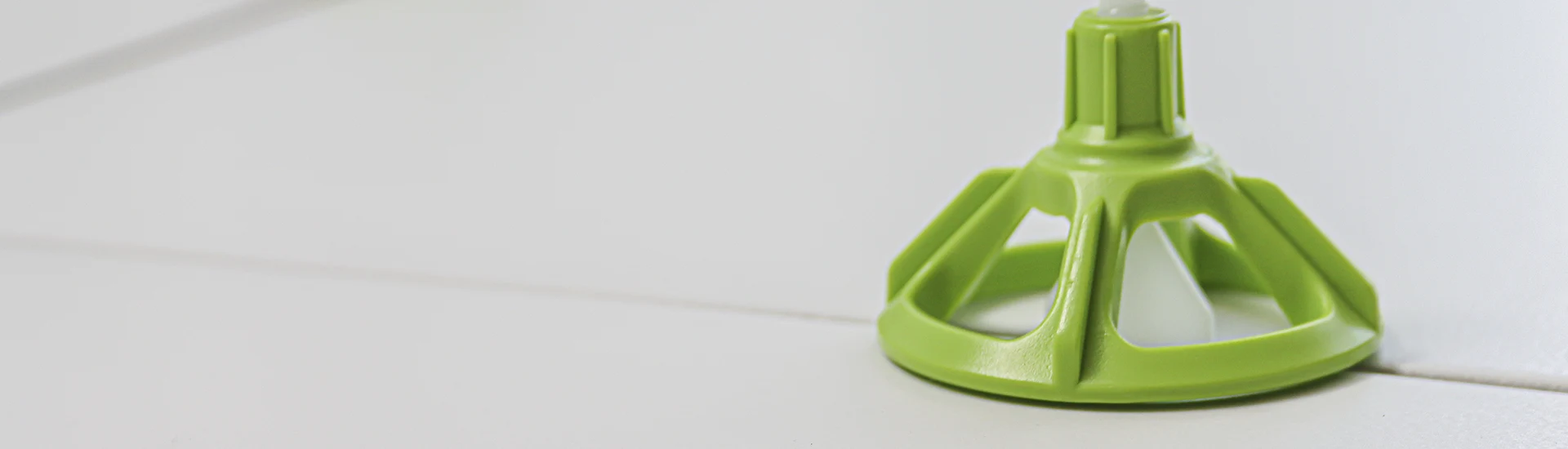 A panoramic view showing a lone green tile leveling device on a white tiled floor, positioned on the right with ample space around it, illustrating the beginning of a tiling project.